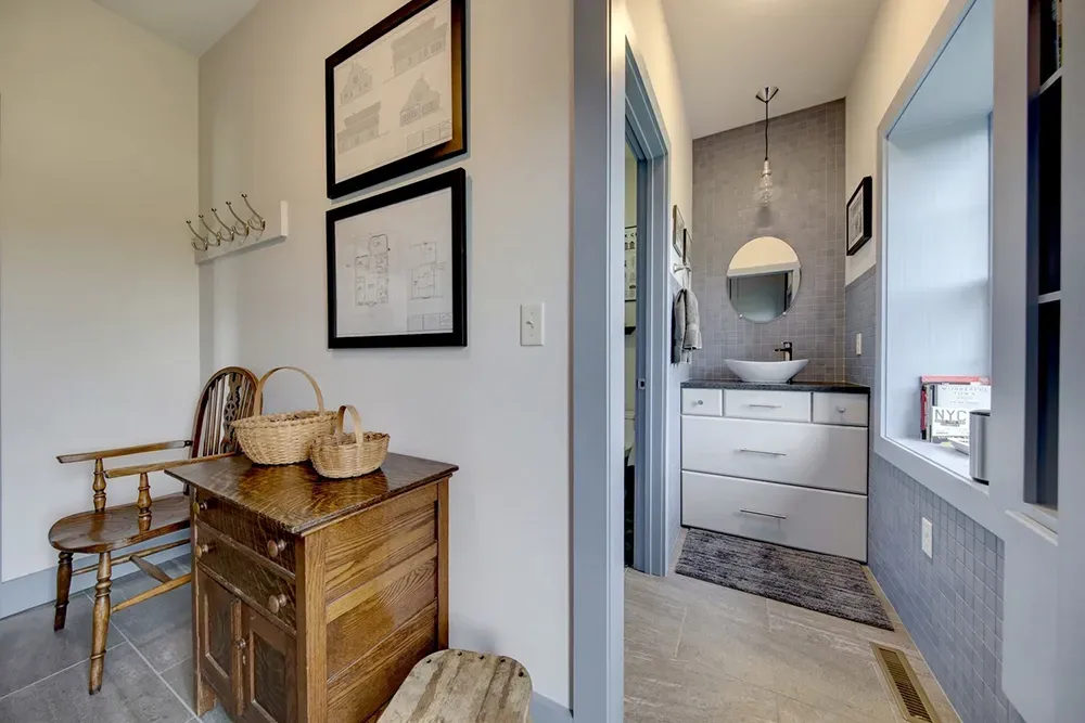 A foyer with a wooden bench and cabinet, leading to a bathroom featuring a white vanity and a round mirror.