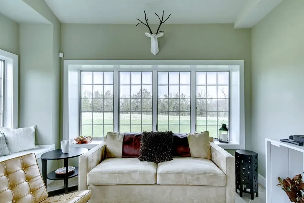 A beige sofa sits before a large window with a white deer head mount above it in a soft-toned, cozy room.
