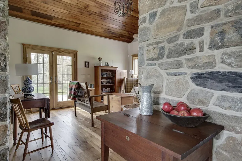 A rustic living room with stone walls, wood floors and ceiling, glass doors, a wooden chair, and a bowl of apples.