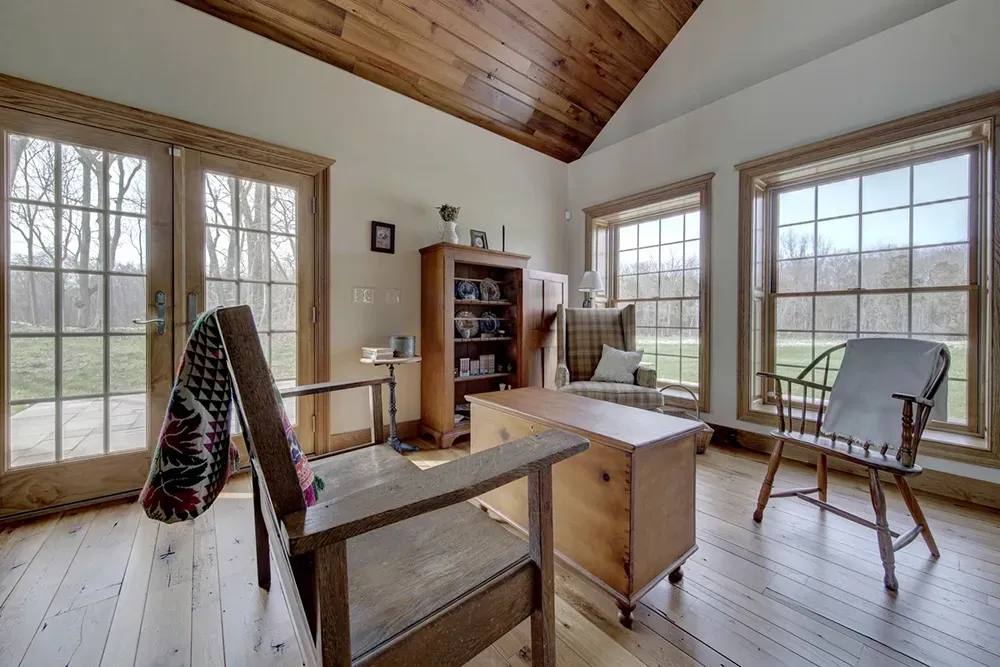 A sunlit room with wooden floors, pine ceiling, two chairs, a small table, and a bookshelf, looking out to trees.