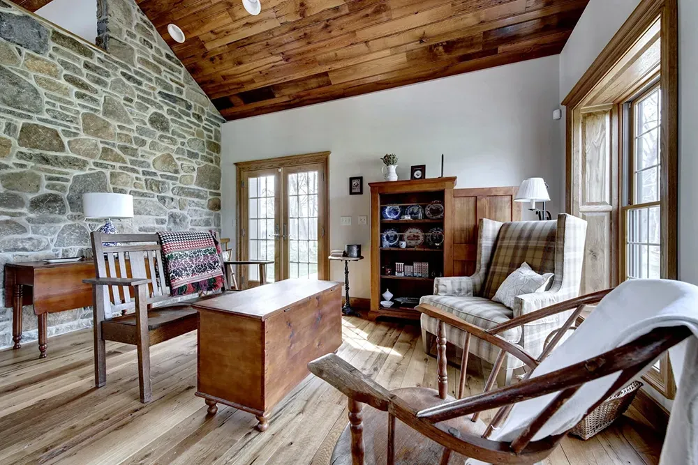 A rustic living room features a stone wall, wood-paneled ceiling, wooden furniture, and a plaid armchair.