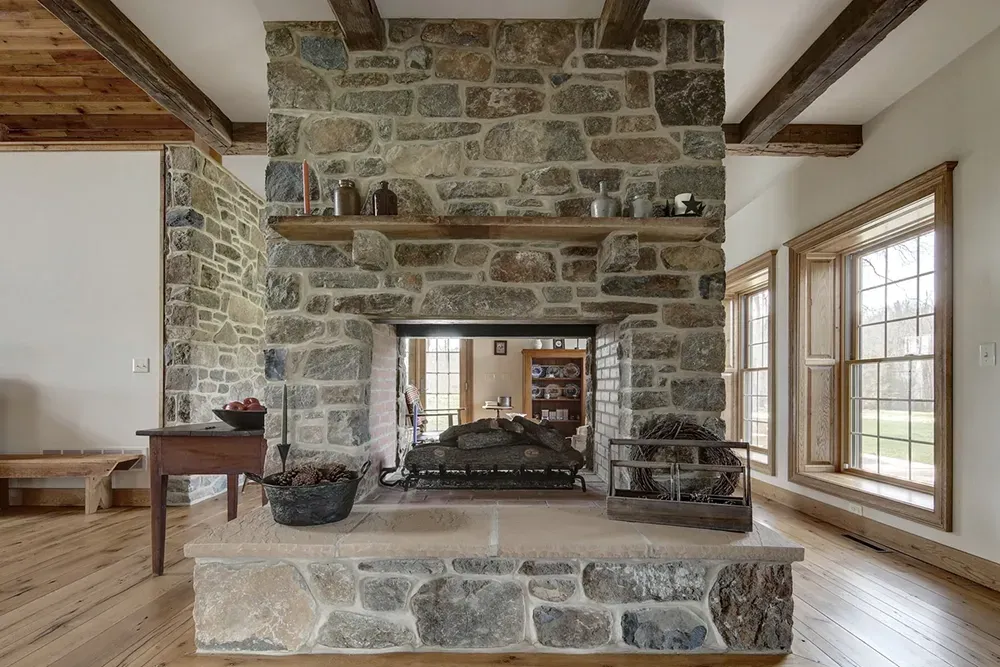 A large, double-sided stone fireplace with a wooden mantel serves as a room divider in a bright, rustic-style home.