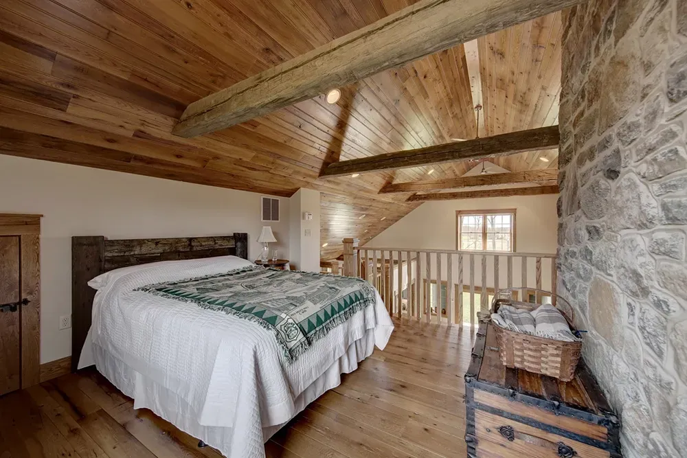 A rustic loft bedroom featuring a bed with a white quilt, wood-paneled ceiling with beams, and a stone accent wall.