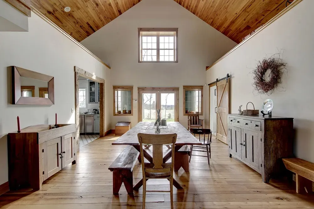 A rustic dining room with a long wooden table, benches, vaulted ceiling, and wooden floors under a central window.