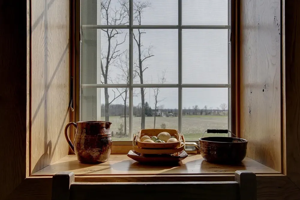 Pottery and a basket of eggs sit on a wooden windowsill overlooking a rural landscape with trees and fields.