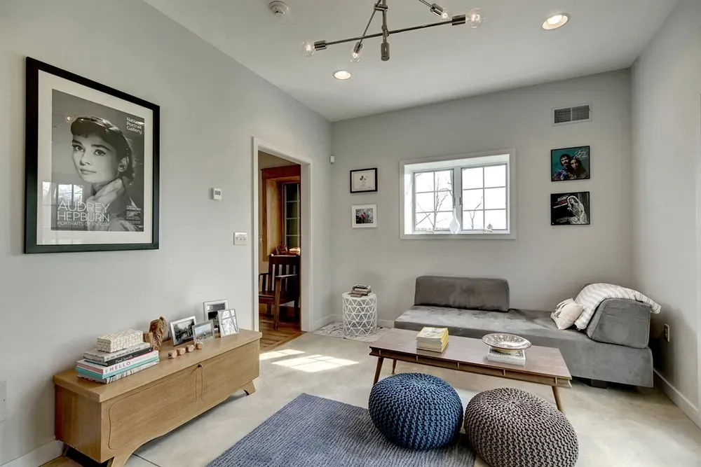 A brightly lit living room with light walls, a wooden console, a grey daybed, two poufs, and framed artwork.