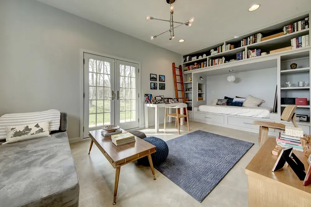 A bright living area featuring a daybed with built-in bookshelves, a wood coffee table, a blue rug, and French doors.