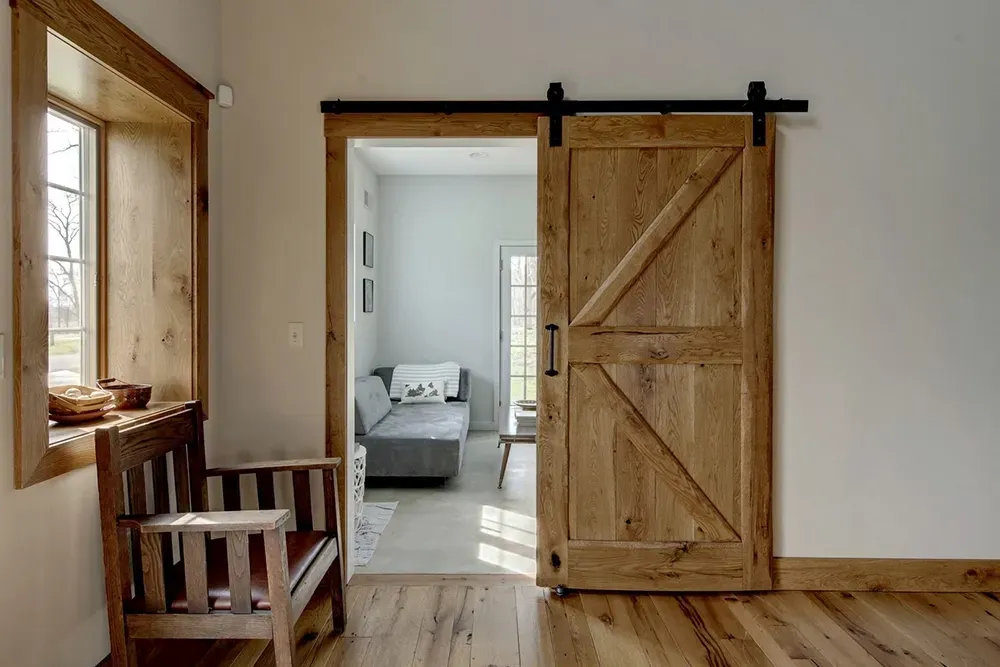 A rustic barn door slides open to a room with a sofa, featuring a wooden chair in the foreground and light wood floors.