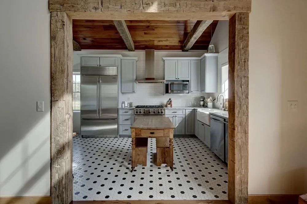 A rustic kitchen framed by reclaimed wood beams, featuring light cabinets, a small wooden island, and black-spotted tile.