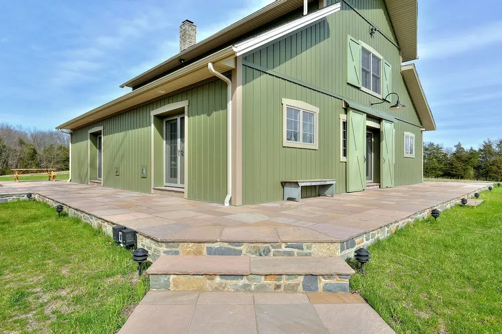 A sage green barn-style house with stone steps leading up to a stone patio under a bright blue sky.