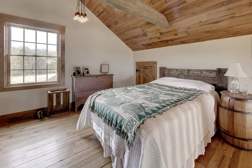 A cozy bedroom with a wooden ceiling, a bed with a green patterned throw, a barrel nightstand, and a window.