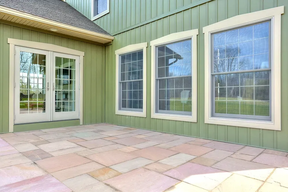 A green house with vertical siding featuring a patio door and three windows, adjacent to a light-colored stone patio.