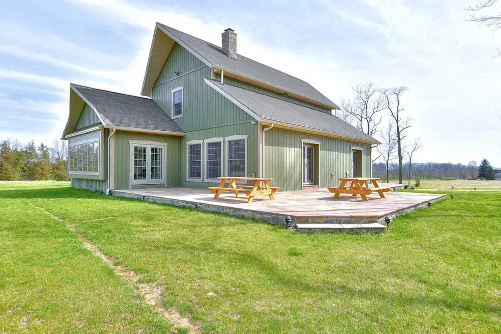 A green, two-story house with a stone patio, two picnic tables, and a large yard under a bright, partly cloudy sky.