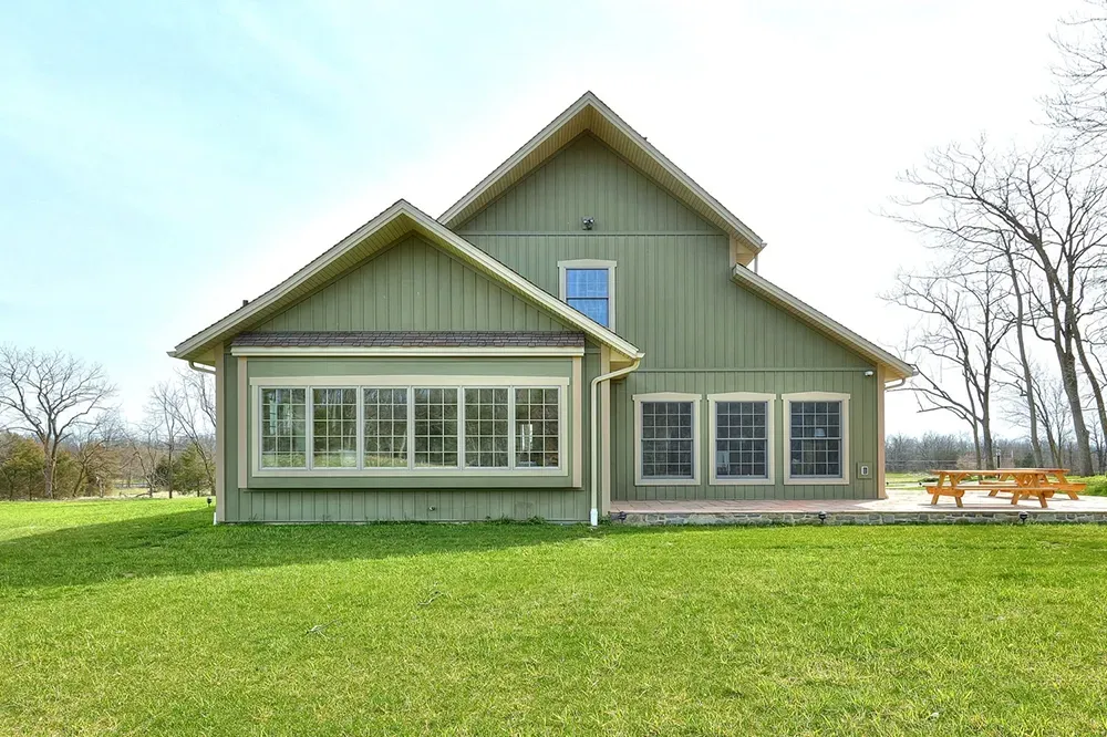 A two-story, olive green house with a prominent multi-pane window and wooden deck on a grassy lawn under a clear sky.