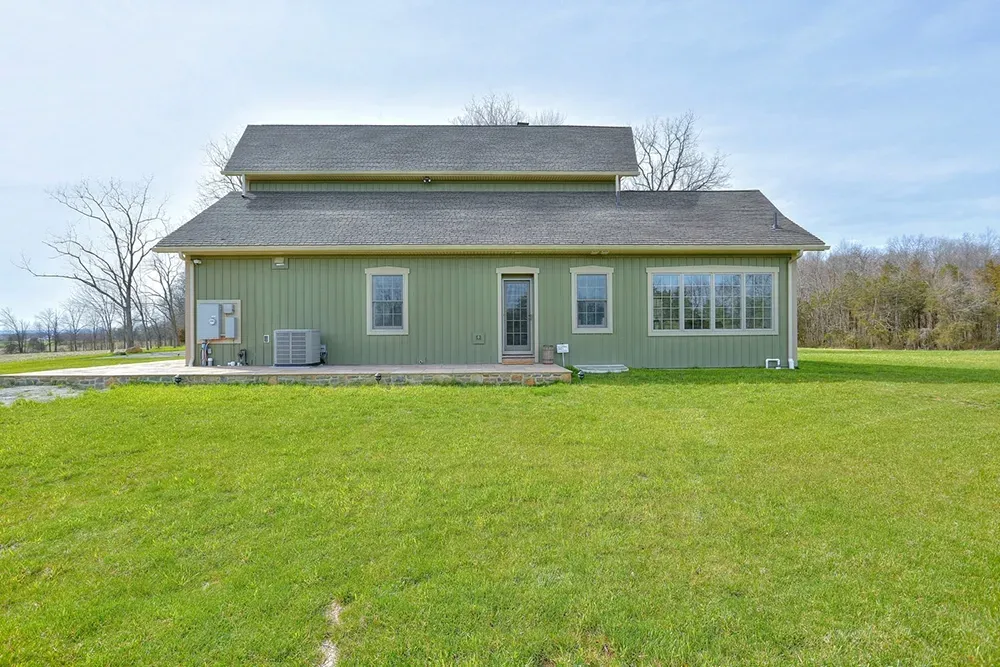 A two-story, olive green house with a dark gray shingled roof, set against a large, grassy lawn under a clear blue sky.
