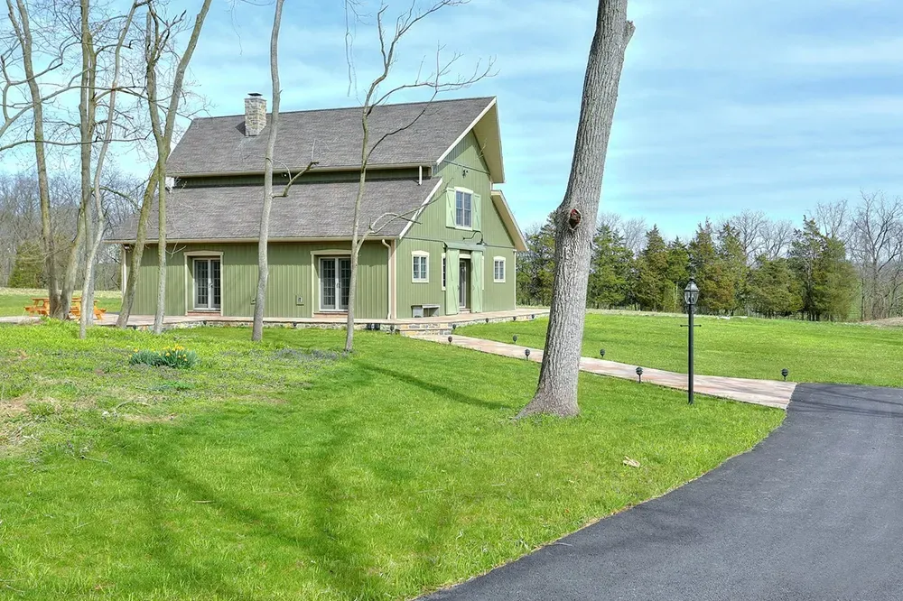 A green barn-style house with a gray roof sits on a grassy lawn under a blue sky, with a paved driveway and stone path.