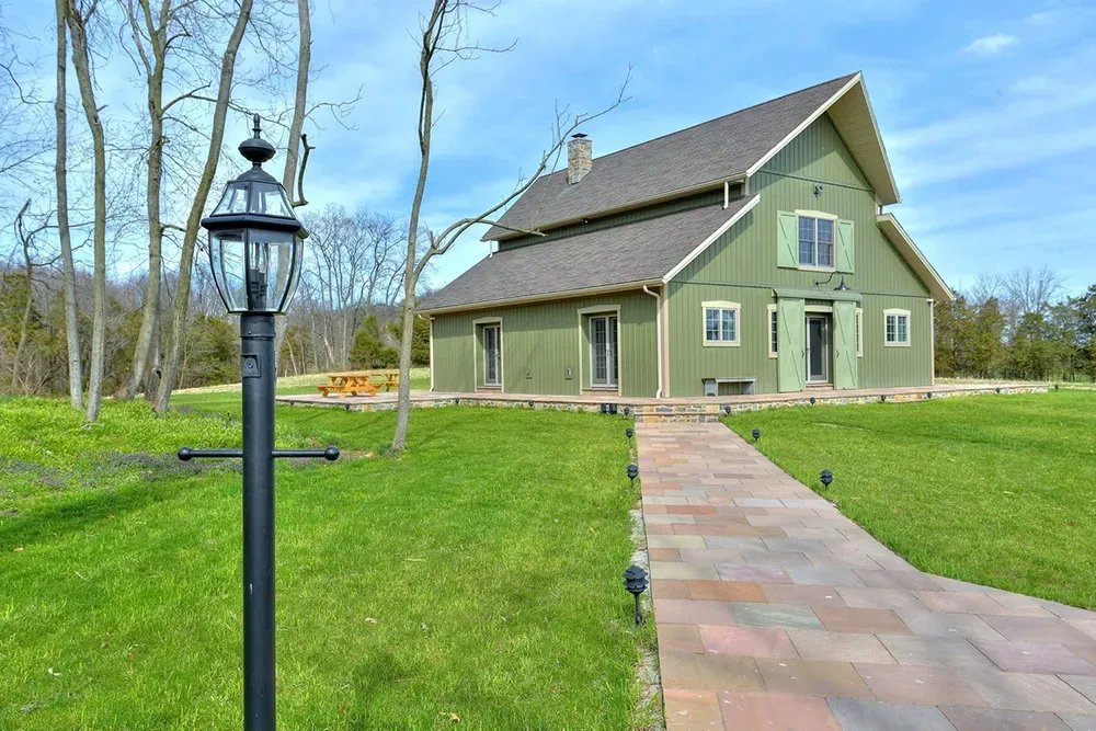A green, two-story house with a shingled roof, set on a grassy lawn with a stone path and a black lamp post in front.