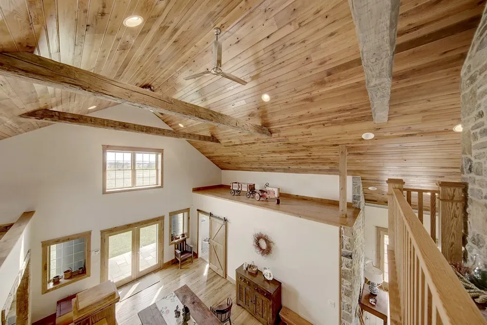 High-angle view of a living room with white walls, wood-paneled vaulted ceilings, exposed beams, and a loft overlook.