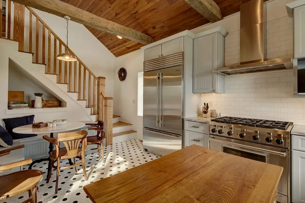 A bright kitchen with a wooden ceiling, a staircase over a dining nook, a stainless steel fridge, and a wooden island.