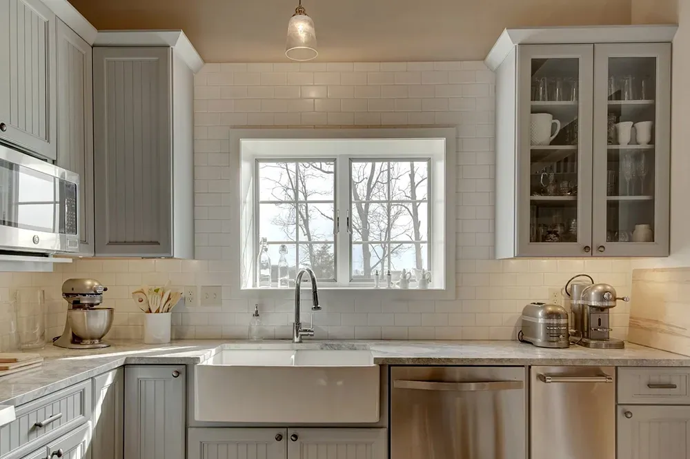 A bright kitchen with white subway tile, a farmhouse sink, stainless appliances, and grey shaker-style cabinets.