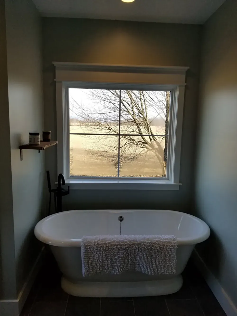 A white freestanding bathtub sits in front of a window overlooking a field, with a small wall shelf to the left.