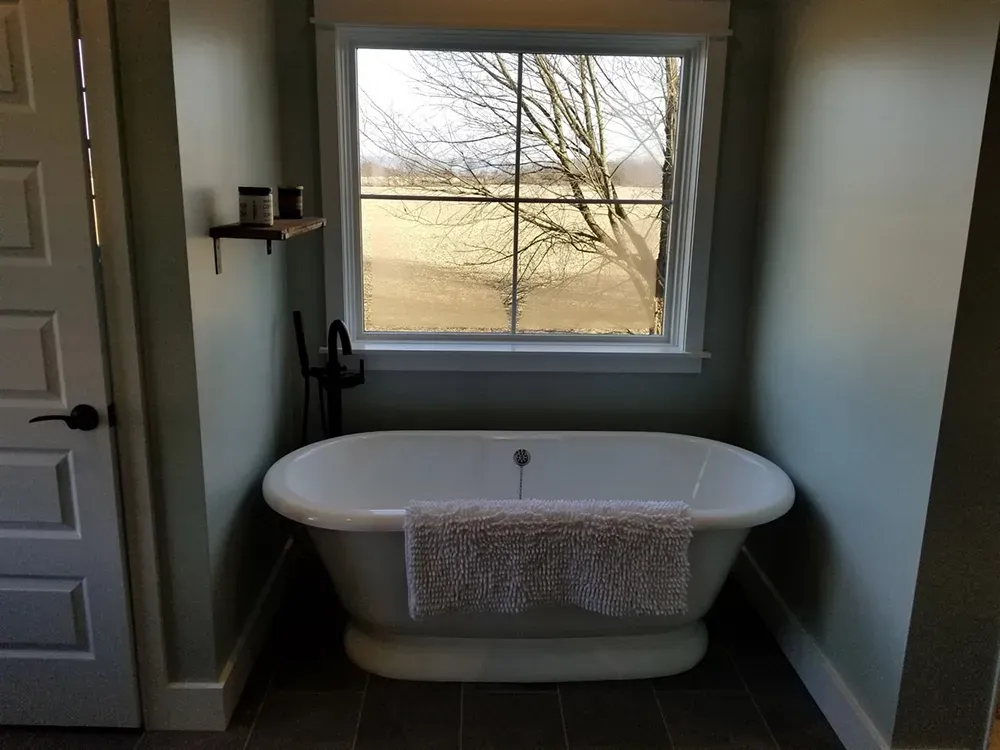 A white freestanding soaking tub sits in a bathroom under a large window overlooking a field, with a rug on the side.
