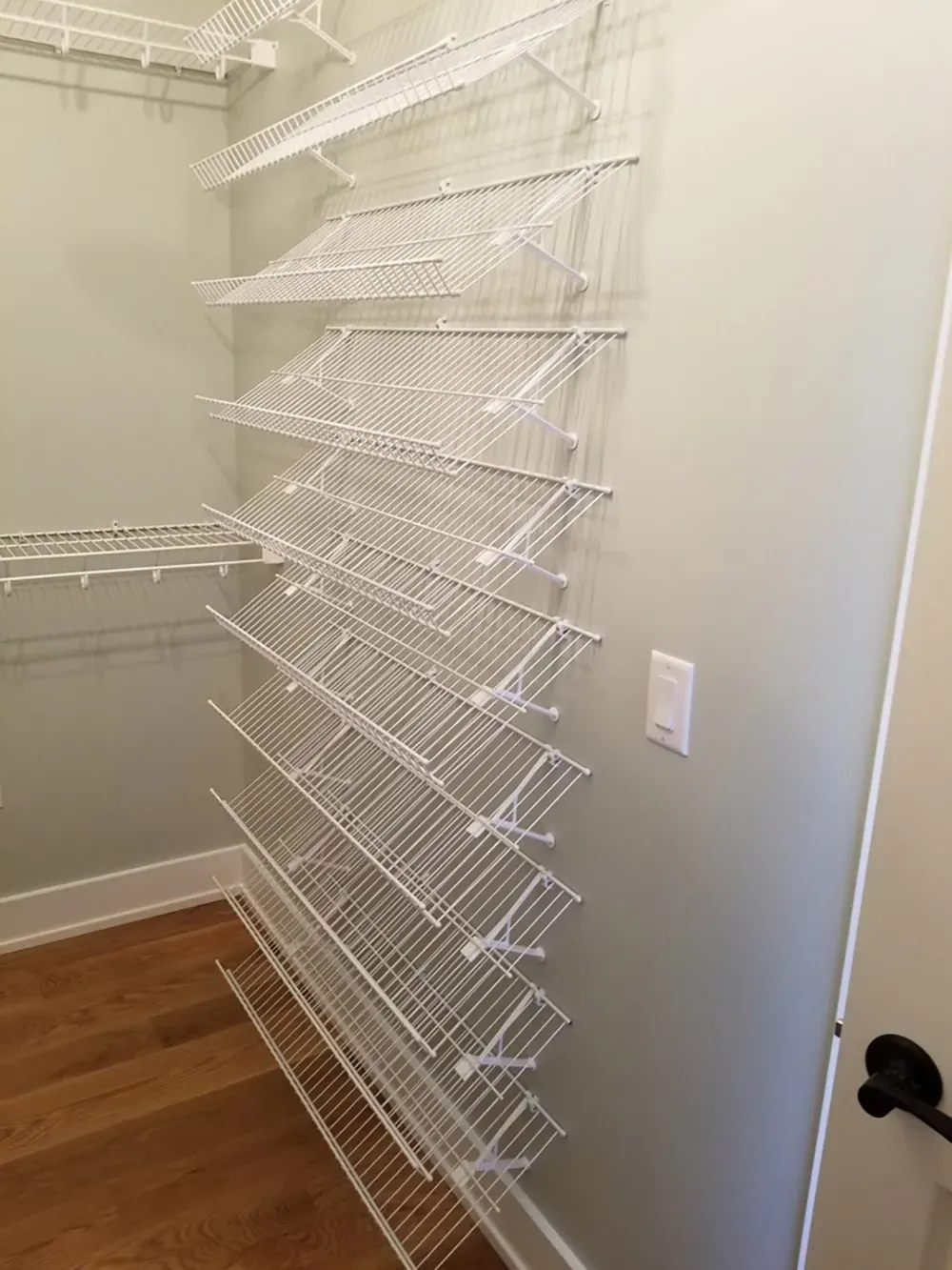 A closet wall with many white wire shoe racks mounted at a slanted angle in a room with light green walls and wood floors.