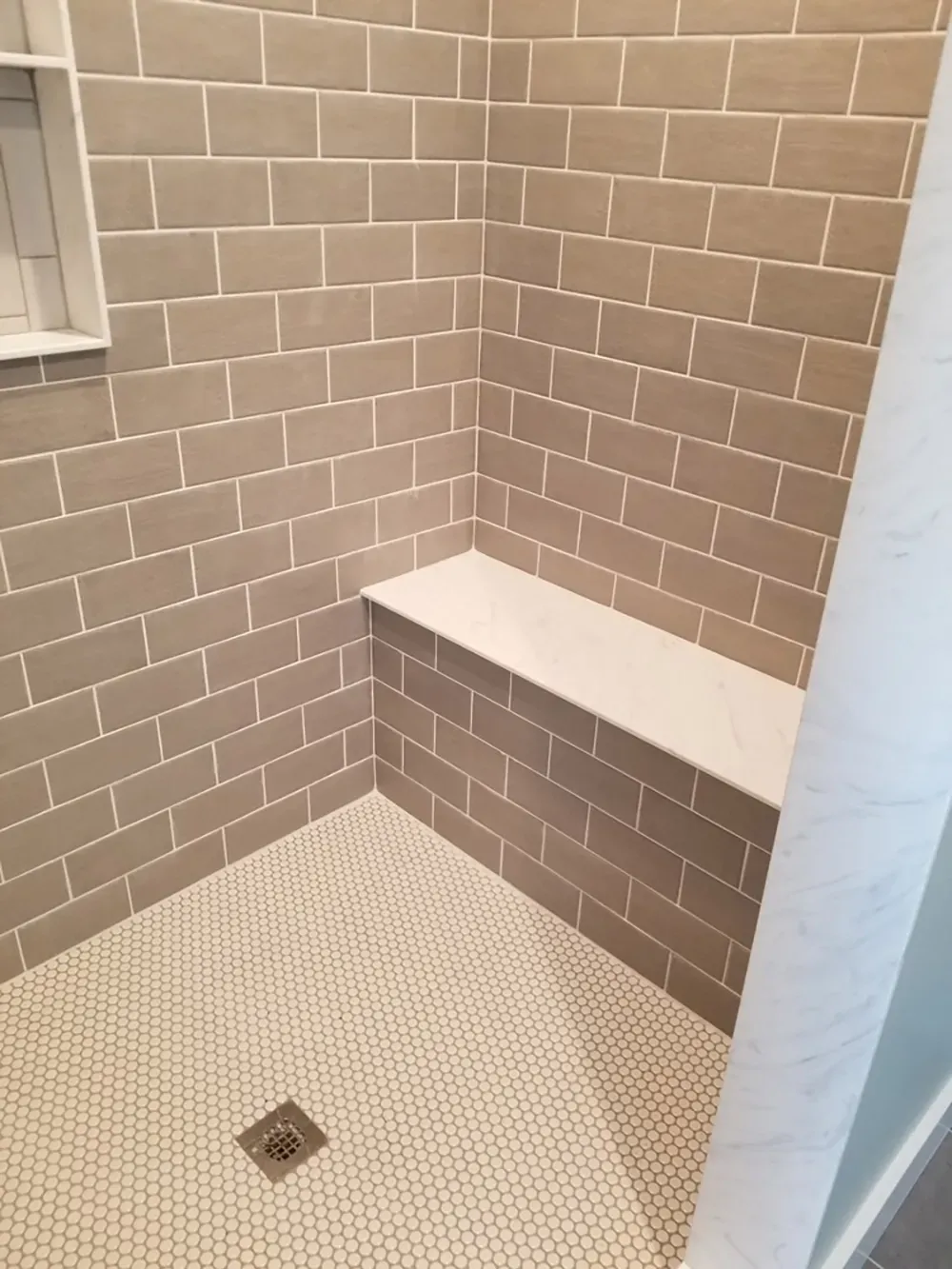 A shower stall with tan subway tile walls, a white stone bench, and small circular white tile flooring.