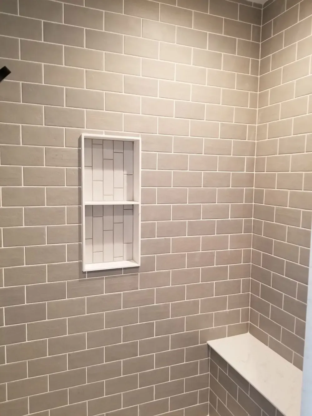 A shower wall featuring gray subway tiles, a white recessed shelf with white vertical tile backing, and a corner bench.