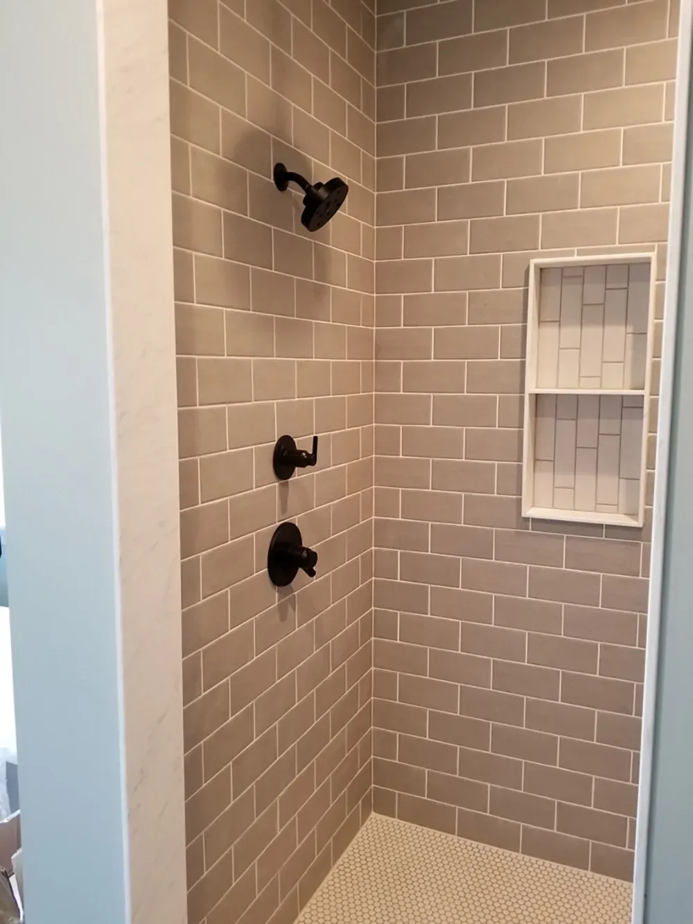 A walk-in shower with gray subway wall tiles, a white recessed niche, a black showerhead and handle, and mosaic floor tiles.