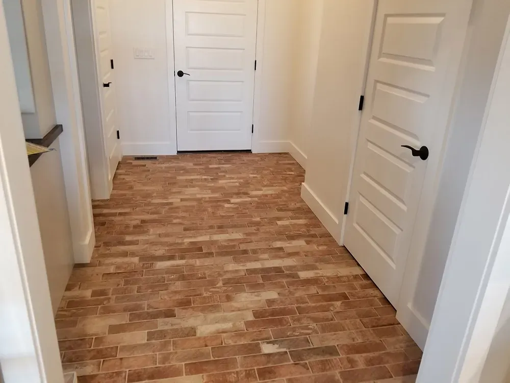 An indoor hallway with light brown brick flooring and white walls, leading to three white paneled doors.