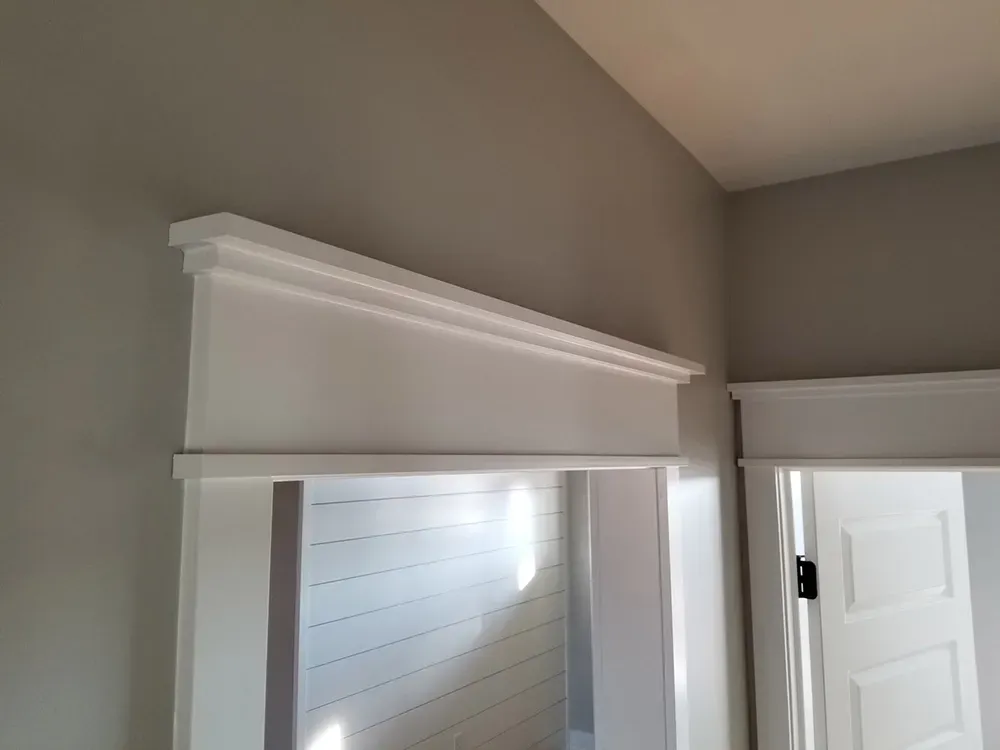 A decorative white wooden header or crown molding installed above a doorway in a room with light gray walls.