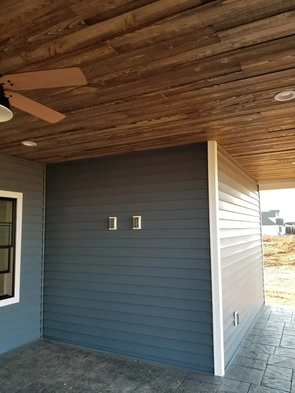 A covered outdoor patio featuring dark blue horizontal siding, a rustic wood plank ceiling, and a ceiling fan.