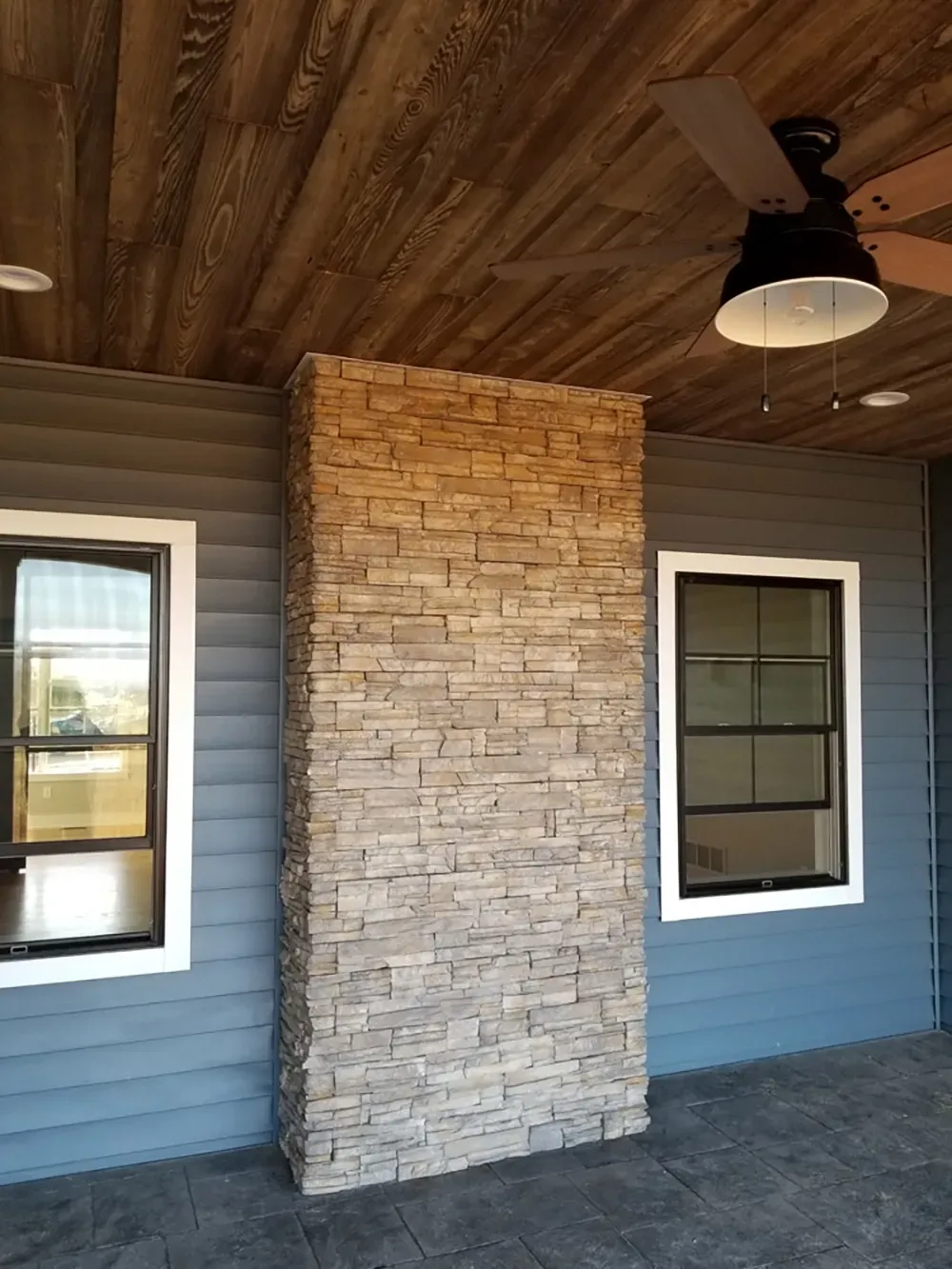 A stone column stands on a covered porch with gray horizontal siding, two white-trimmed windows, and a wooden ceiling.