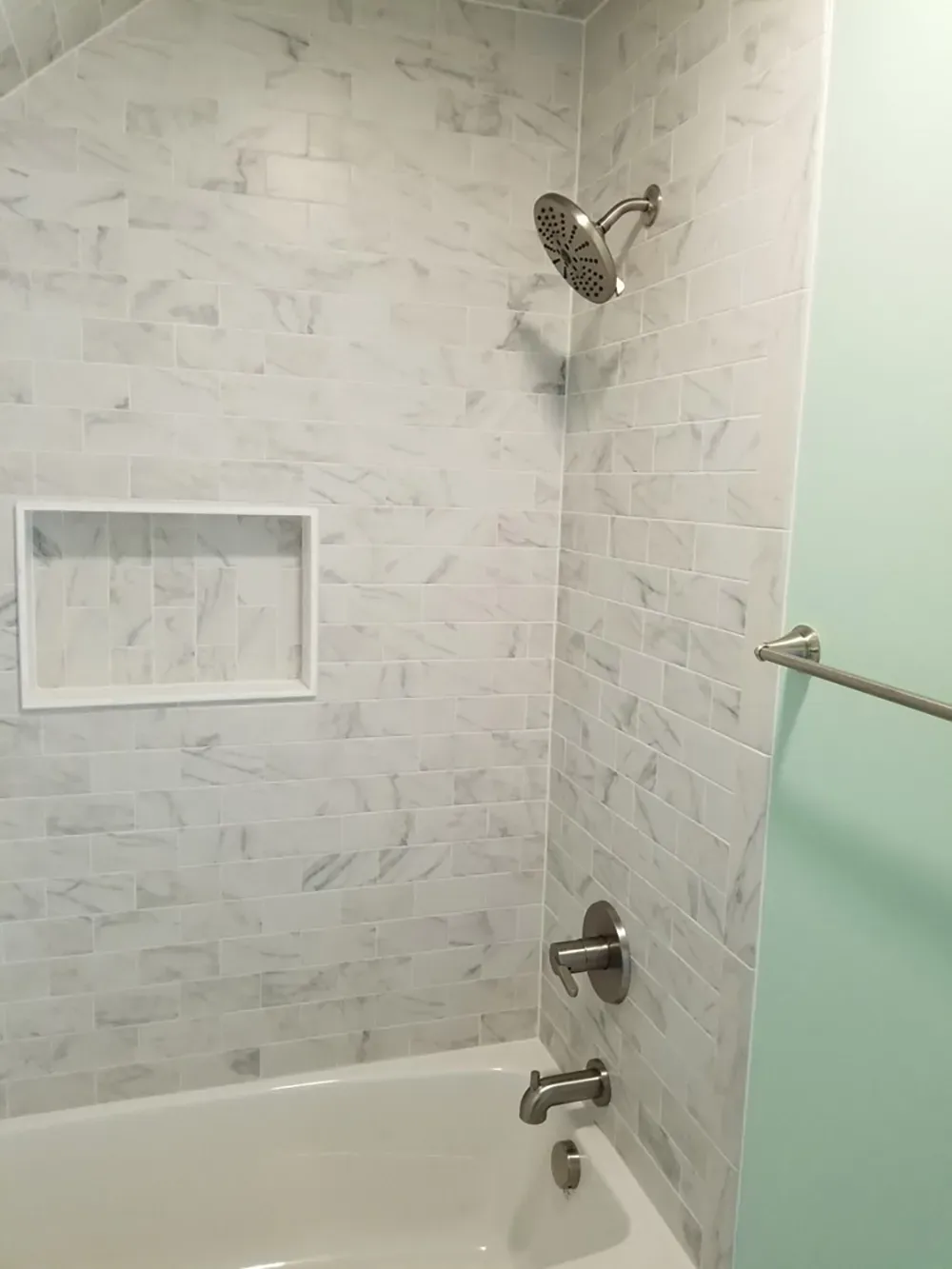 A shower area with white marble-style tiled walls, a built-in recessed shelf, a bathtub, and brushed metal fixtures.