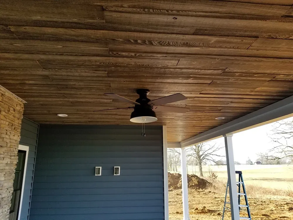 A dark wood-paneled porch ceiling with a central black ceiling fan, viewed against blue horizontal siding and a field.