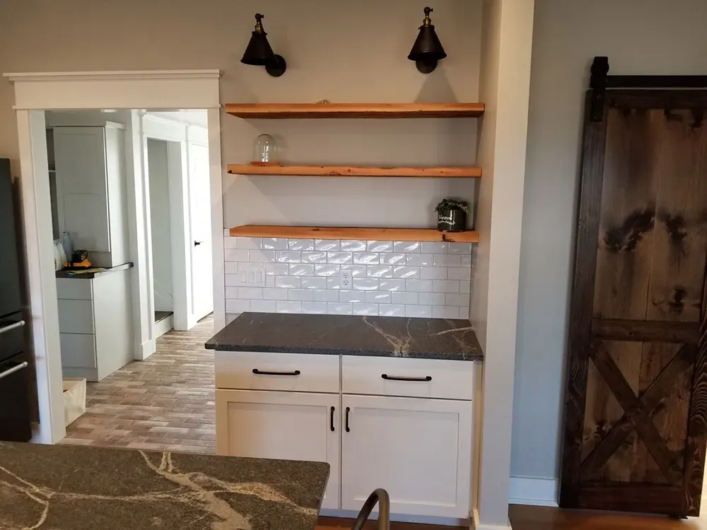 White kitchen cabinetry with black stone counters, three wood floating shelves, and two wall lamps on a tiled backsplash.