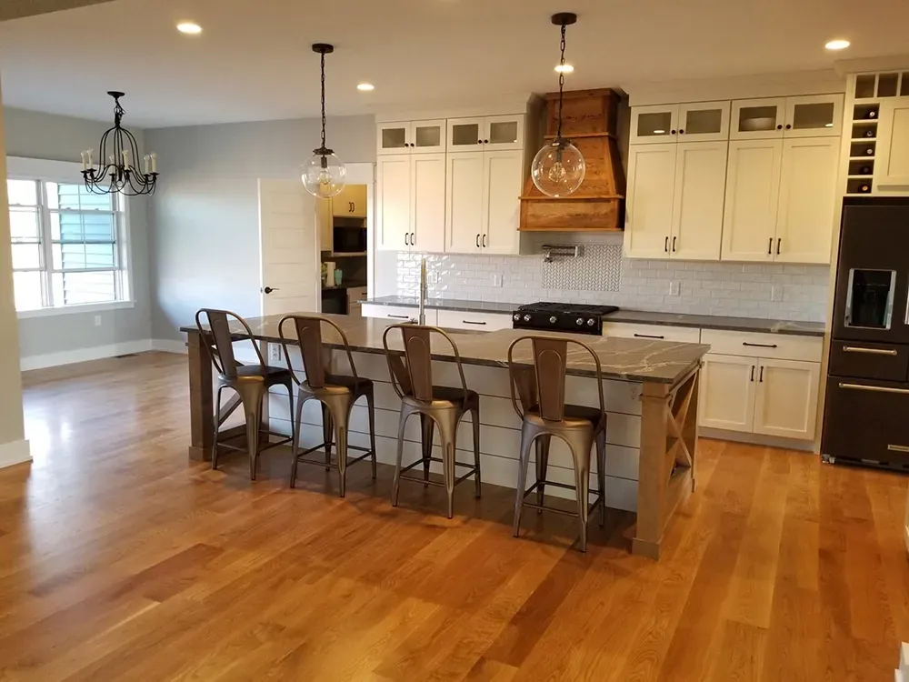 A kitchen with white cabinets, a wood range hood, a large island with four metal stools, and hardwood floors.