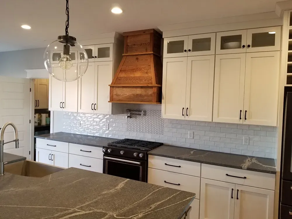A modern kitchen with white cabinets, dark stone countertops, a wood range hood, and a glass pendant light.