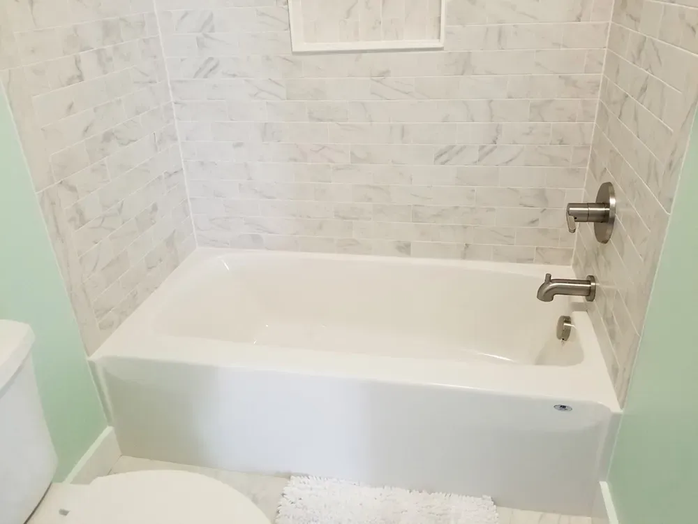 A white bathtub with marble-patterned tile walls and nickel fixtures in a bathroom with light green walls.