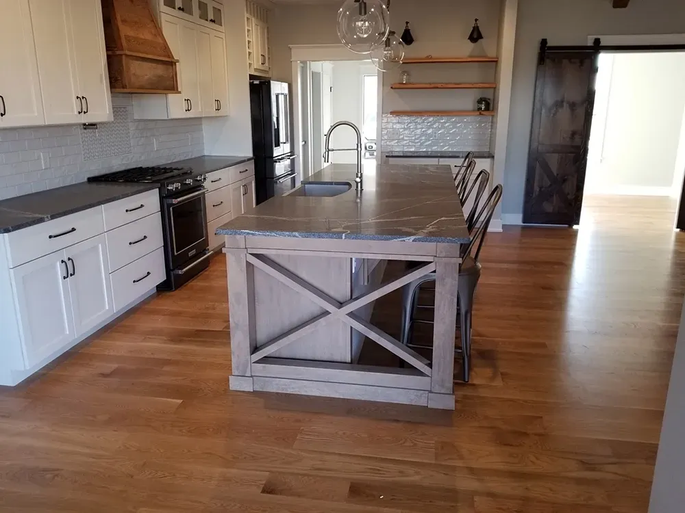 A modern kitchen with white cabinets, a large farmhouse-style island, dark countertops, and light wood flooring.