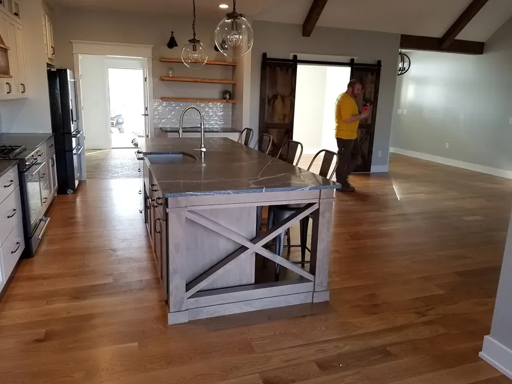 A kitchen with a large wooden island, stainless appliances, open shelving, and light wood floors, with a person standing.