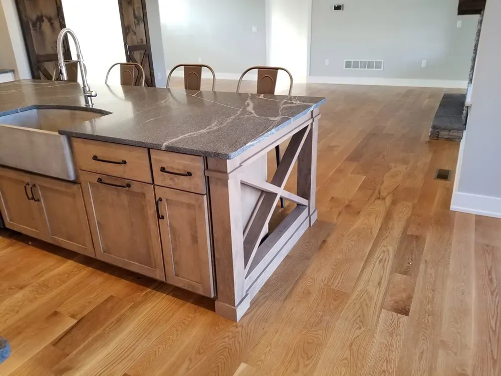 A kitchen island with a farmhouse sink and X-brace detail, featuring light wood cabinets, stone counters, and wood floors.