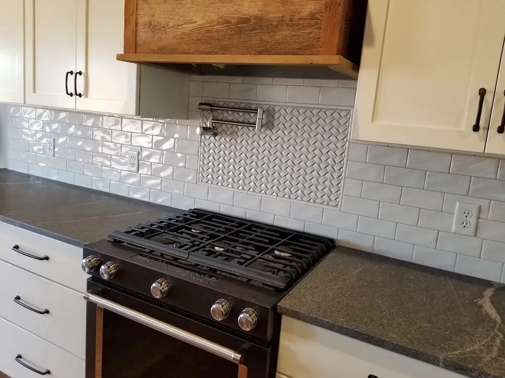 A kitchen counter with a black gas range, white cabinets, grey tile backsplash, and a rustic wooden stove hood.