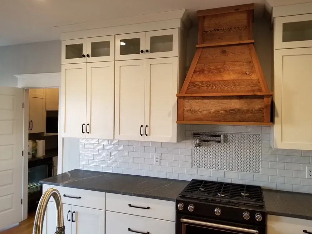 A kitchen featuring white cabinets, a dark countertop, a white subway tile backsplash, and a rustic wooden range hood.