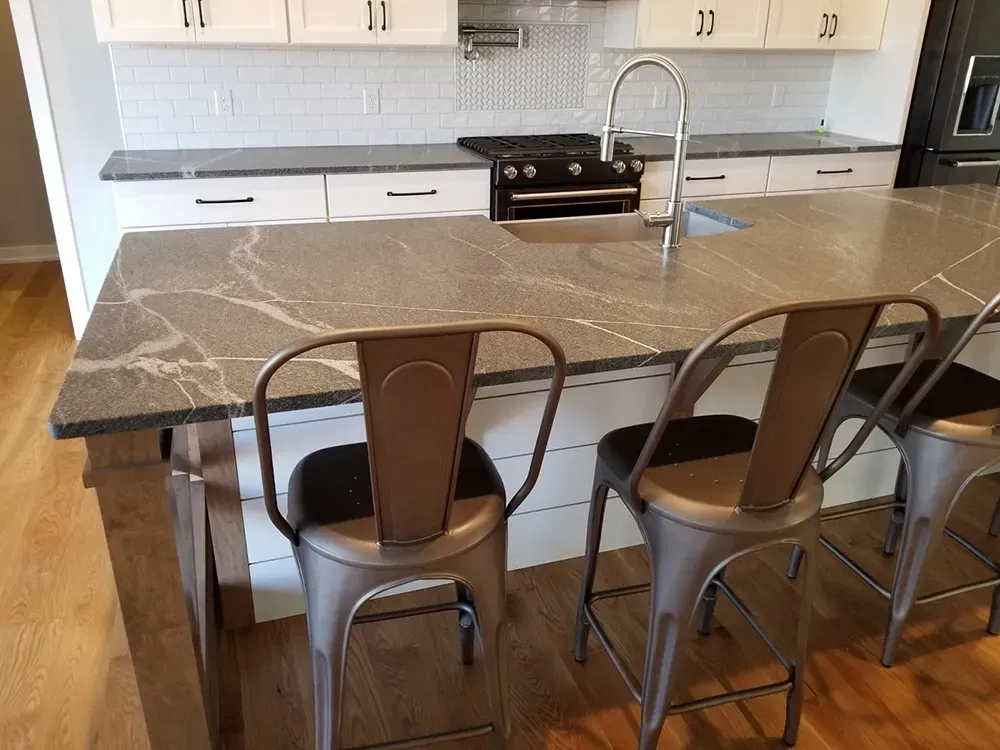 A kitchen island with a dark stone countertop and three metal bar stools in front of white cabinets and a stovetop.