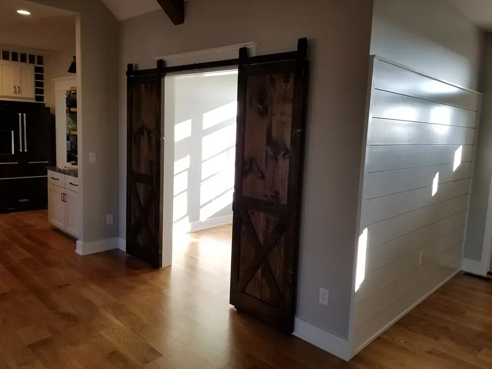 Double dark-stained wooden barn doors with X-pattern accents installed in a white-walled room with hardwood floors.