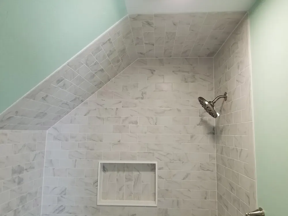 A walk-in shower with light grey marble-style tiled walls, a built-in recessed shelf, and a nickel showerhead.