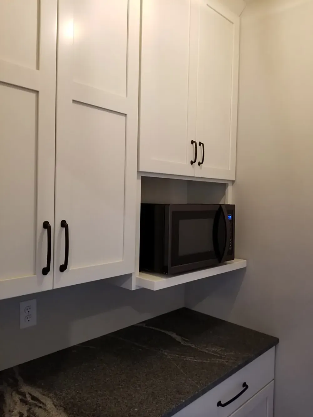 A microwave on a shelf built between white cabinets above a dark stone kitchen counter.