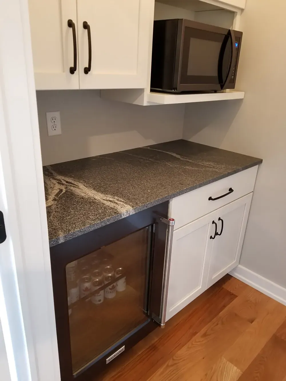 A kitchen bar area with white cabinets, a microwave, granite countertop, and a glass-front beverage refrigerator.